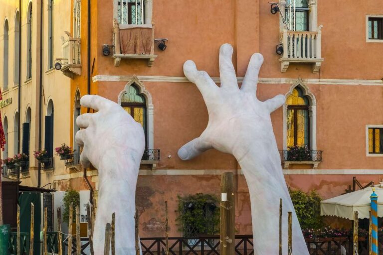 Giant white hand sculpture emerging from a canal to support a building in Venice