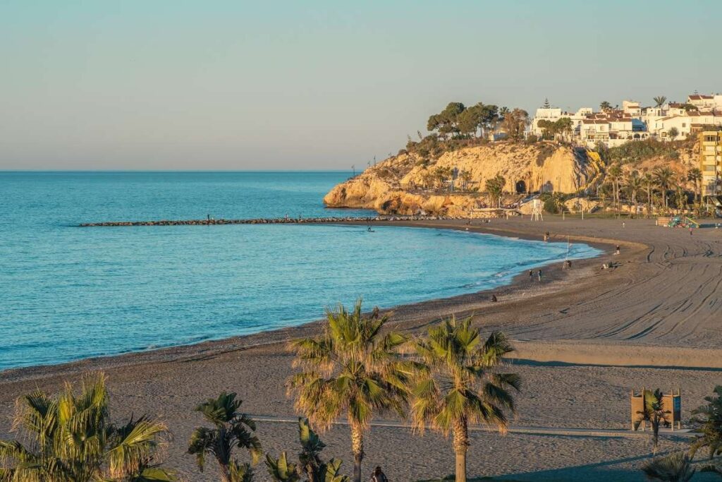 Sunset view of Malaga beach with palm trees and a rocky promontory