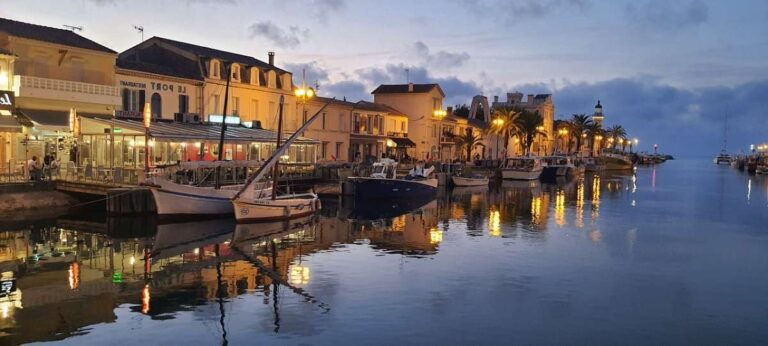 Boats docked in a calm harbor with lit-up buildings at dusk