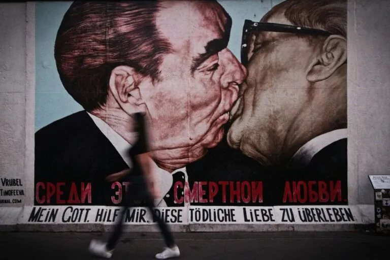 A mural depicting a famous kiss on a Berlin Wall segment with a passerby